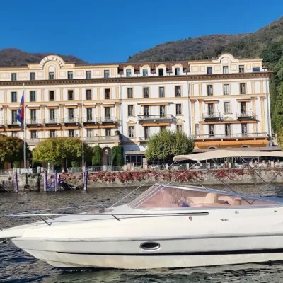 White boat on a lake near a grand hotel with trees and hills in the background.