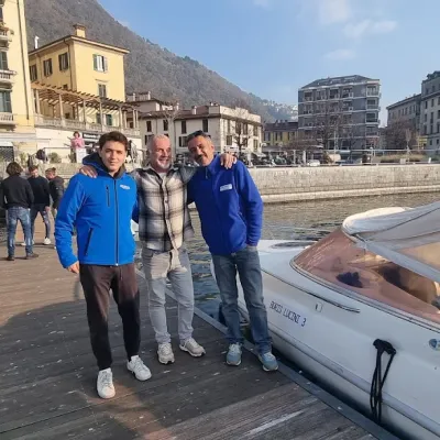 Three men stand on a dock beside two boats, with buildings and a mountain in the background.