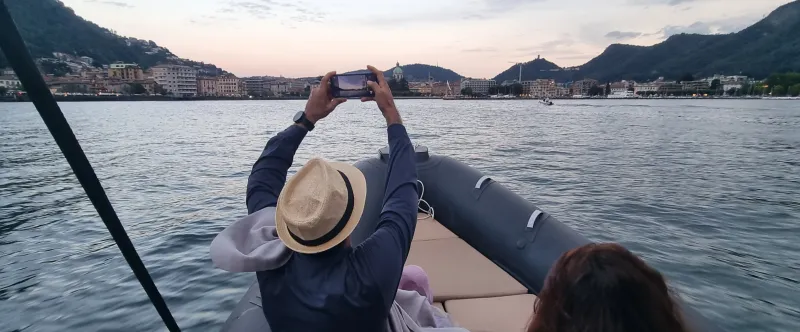 Two people on a boat, one taking a photo of the distant city skyline with hills at sunset.