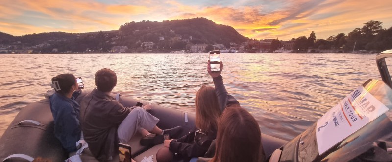 People on a boat at sunset, taking photos on a lake with hills and buildings in the background.