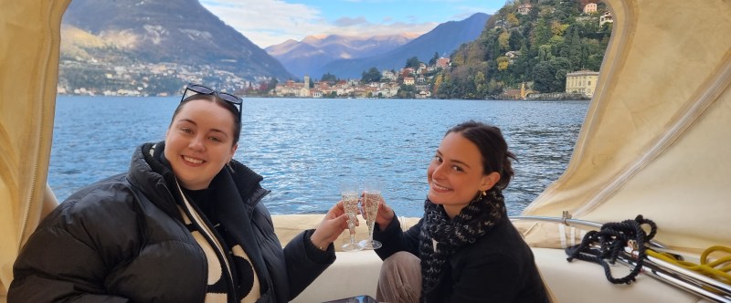 Two people smiling, toasting with drinks on a boat with lake and mountains in the background.
