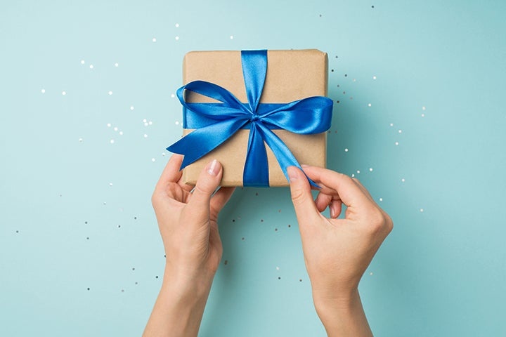 Hands tying a blue ribbon on a gift box against a blue background with confetti.