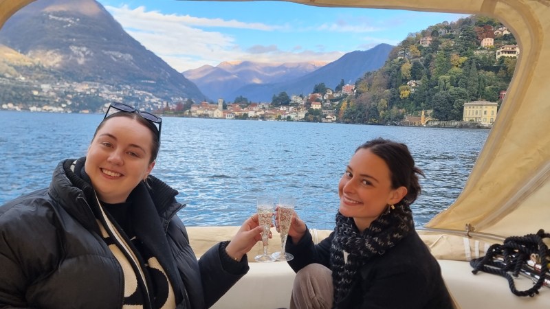 Two people toasting with drinks inside a boat, with scenic lake and mountains in the background.