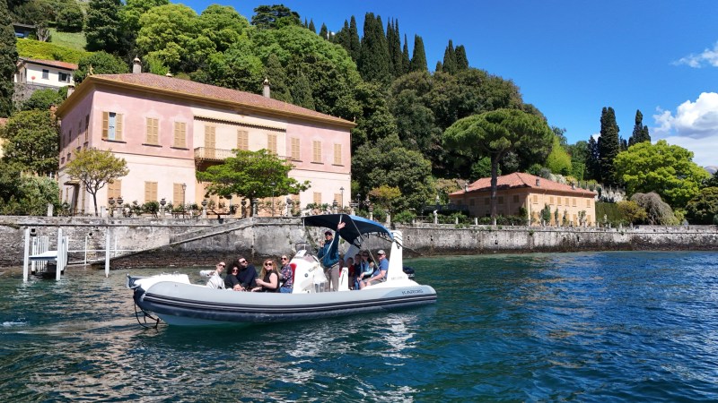 People on a motorboat on a lake with pink villa and trees in the background.