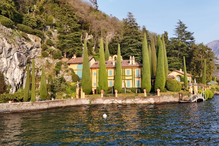 Villa with tall cypress trees by a lakeside, surrounded by cliffs and forested hills.