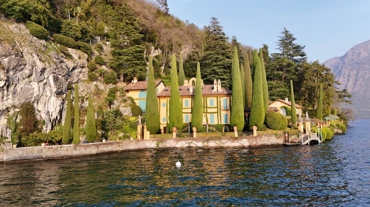Villa with tall cypress trees by a lakeside, surrounded by cliffs and forested hills.