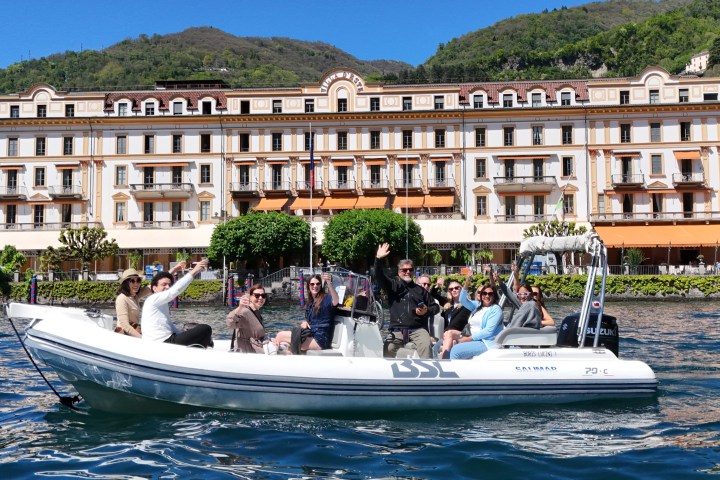 People on a white boat wave, with a large building and hills in the background.