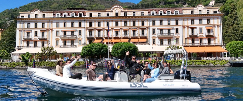 People on a white boat wave, with a large building and hills in the background.