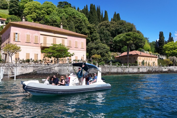 People on a boat in front of a pink villa with green trees and clear blue sky.