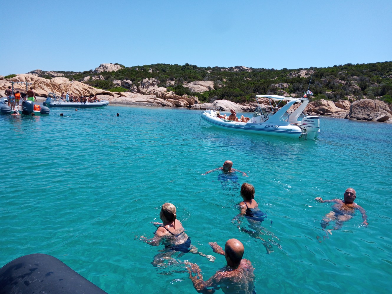 People swimming near boats in clear blue water with rocky shoreline in the background.