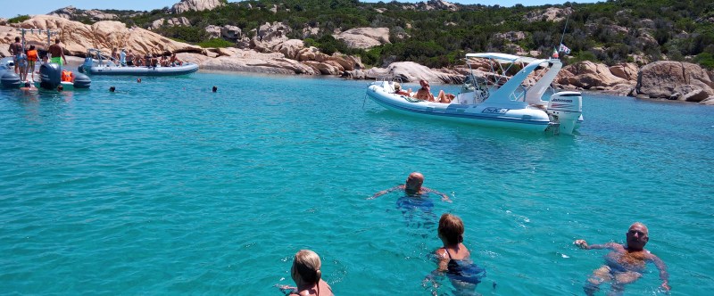People swimming near boats in clear blue water with rocky shoreline in the background.