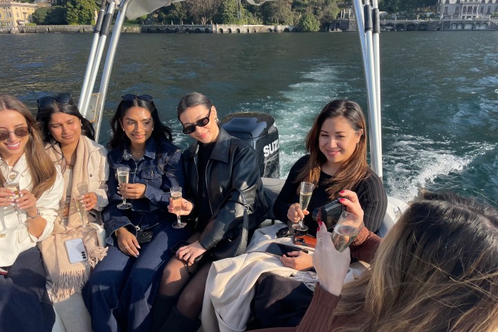 Five people on a boat holding champagne glasses, with water and buildings in the background.