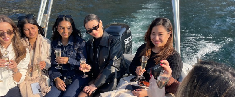Five people on a boat holding champagne glasses, with water and buildings in the background.
