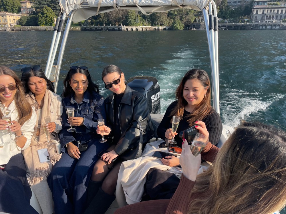 Five people on a boat holding champagne glasses, with water and buildings in the background.