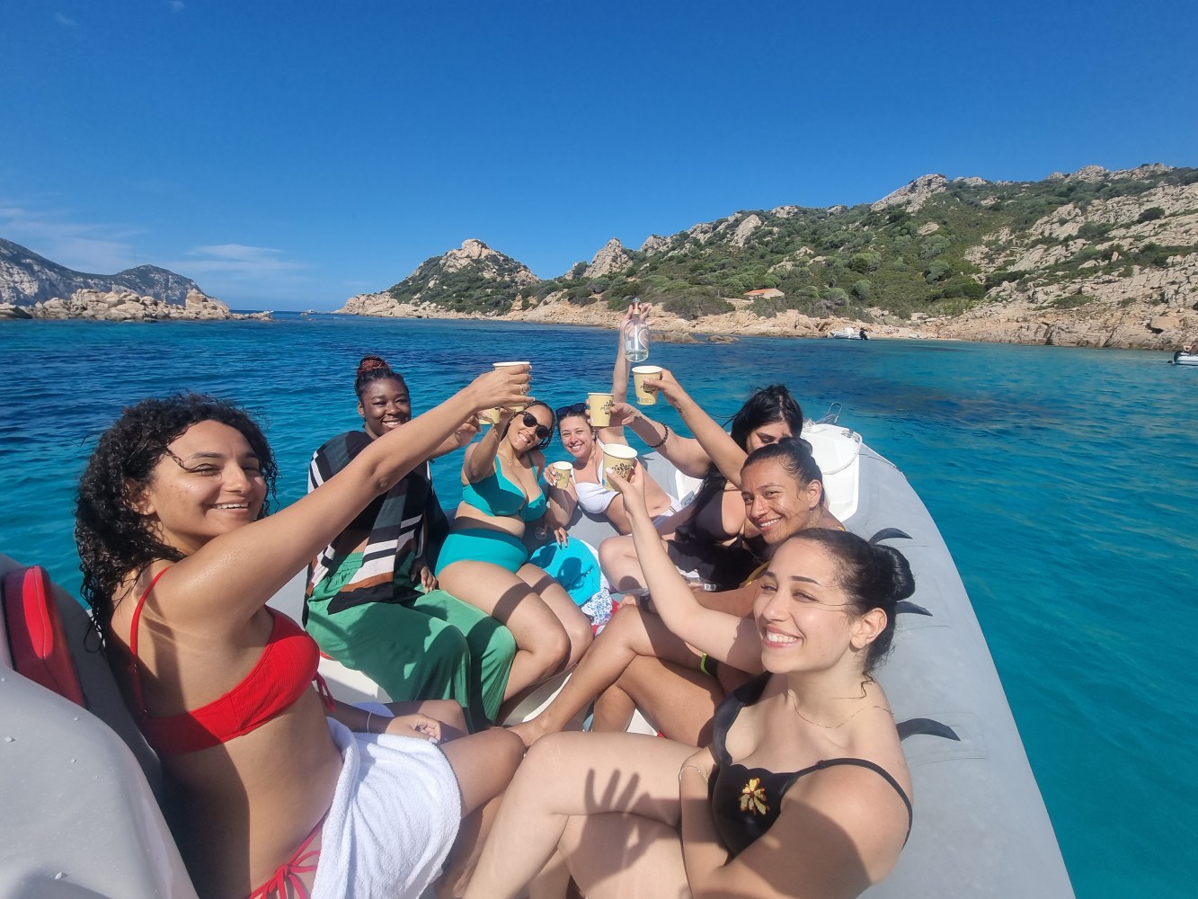 Group of women toasting on a boat in clear blue water near rocky coastline.