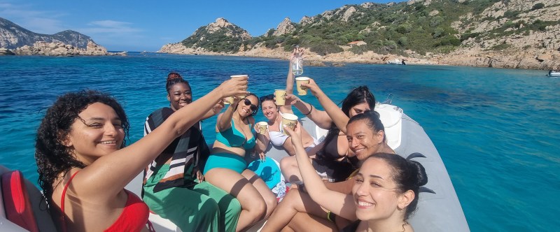 Group of women toasting on a boat in clear blue water near rocky coastline.