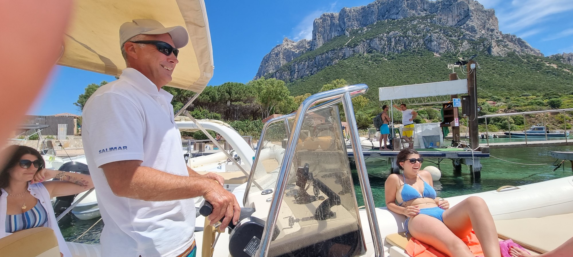 People on a boat with a mountainous backdrop on a sunny day, one person steering, another lounging in swimwear.