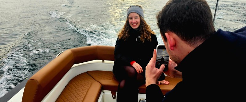 Person photographs woman sitting on a boat with scenic hills in the background.