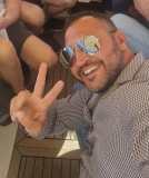 Man in sunglasses and patterned shirt smiling, making a peace sign on a boat with others.