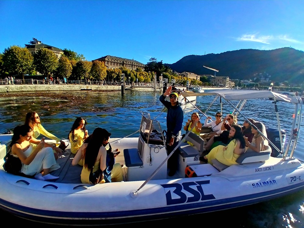 Group of people in a boat on a sunny day with mountains and buildings in the background.