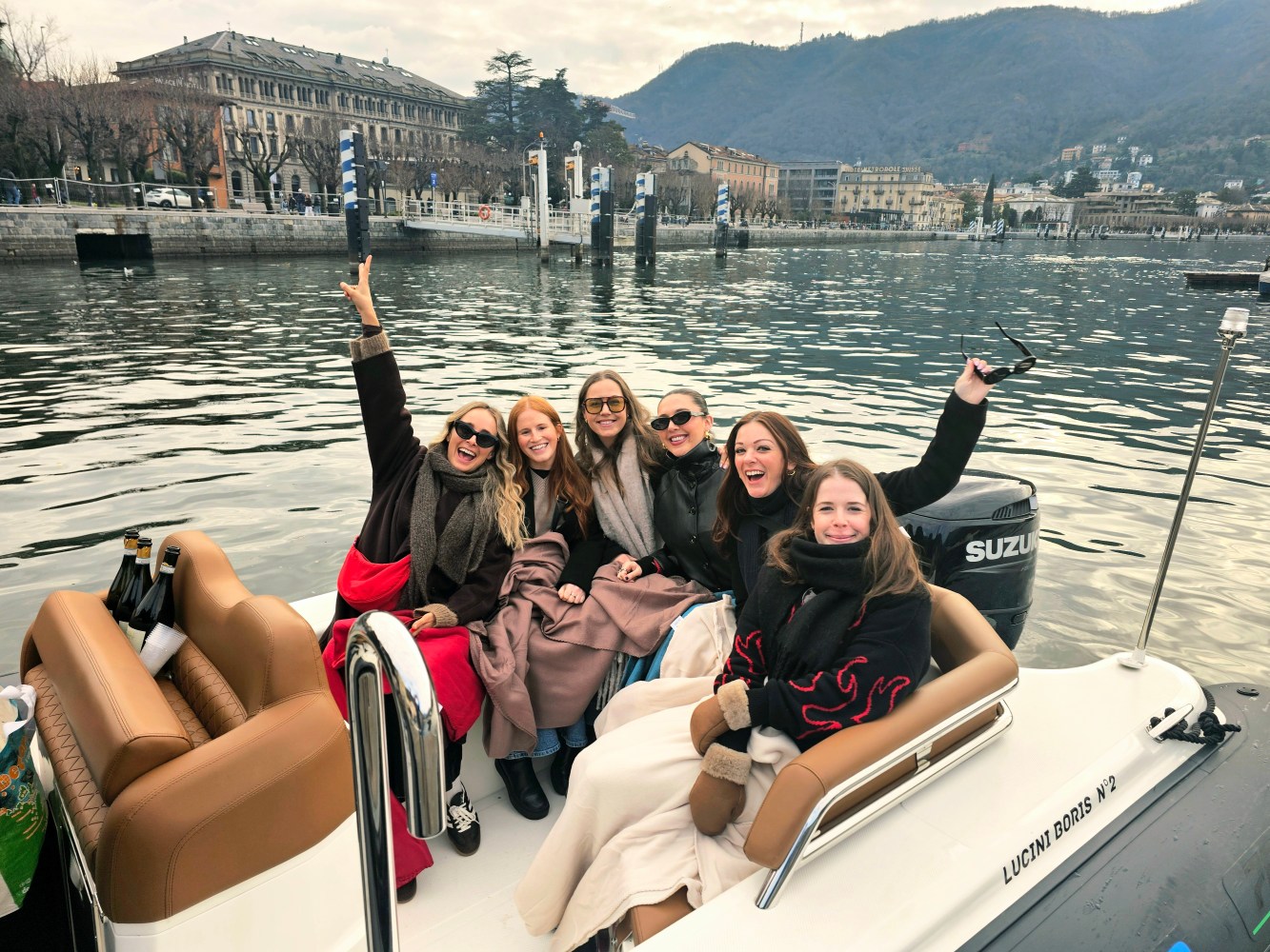 Group of six people smiling on a small motorboat on a lake with buildings and mountains in the background.