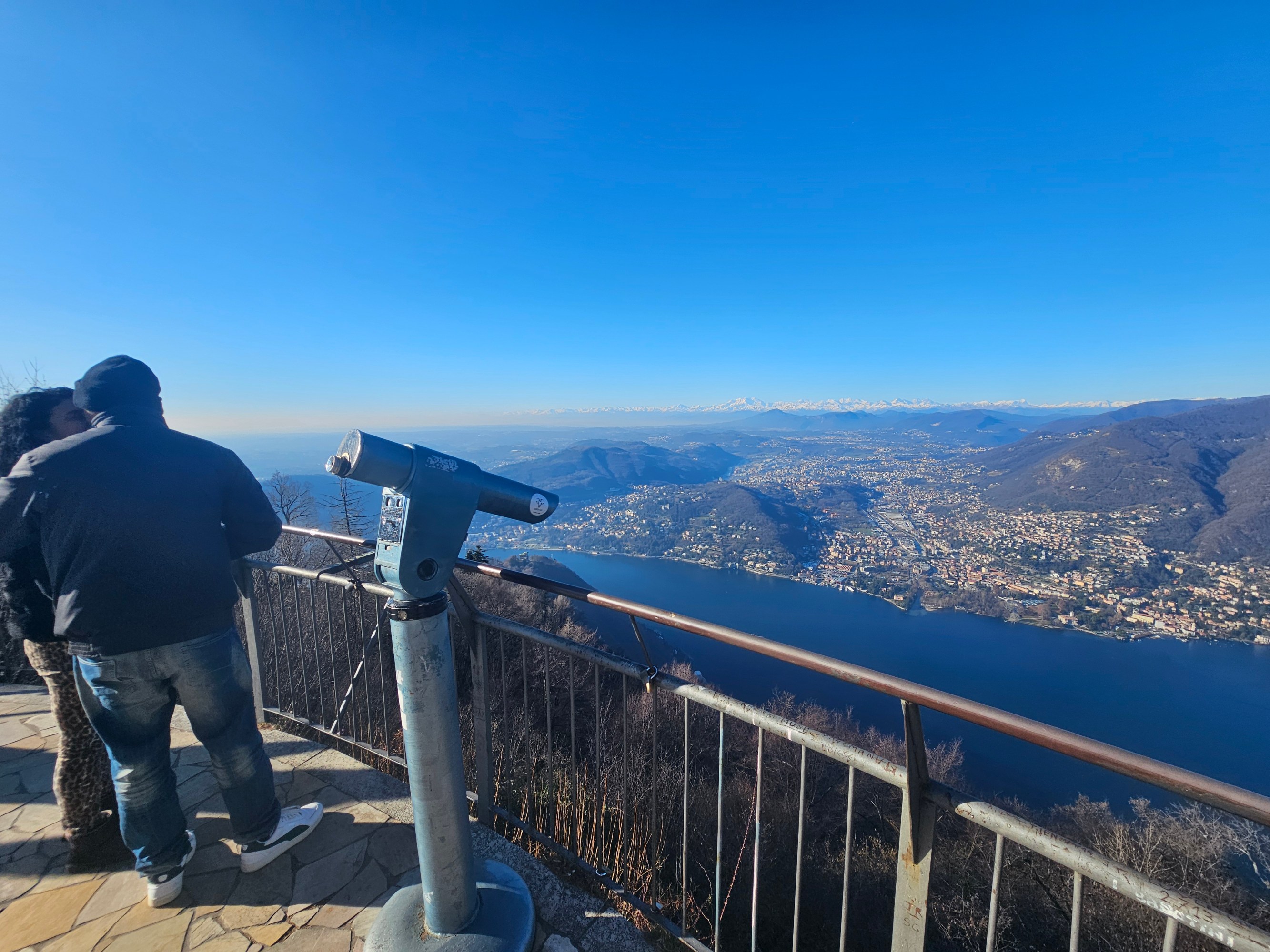 People at a scenic overlook with a telescope, viewing a large lake and distant mountains under a clear blue sky.
