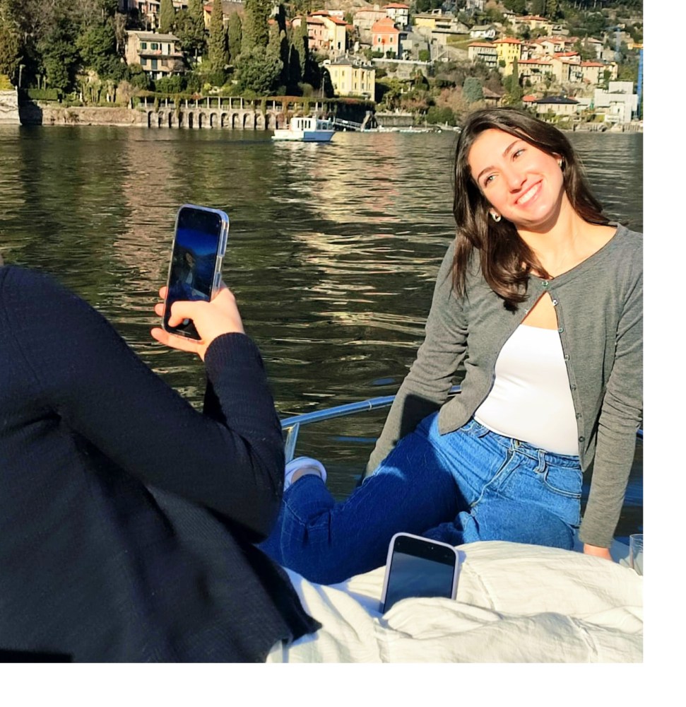 Woman on a boat smiling for a photo, scenic lakeside town in the background.