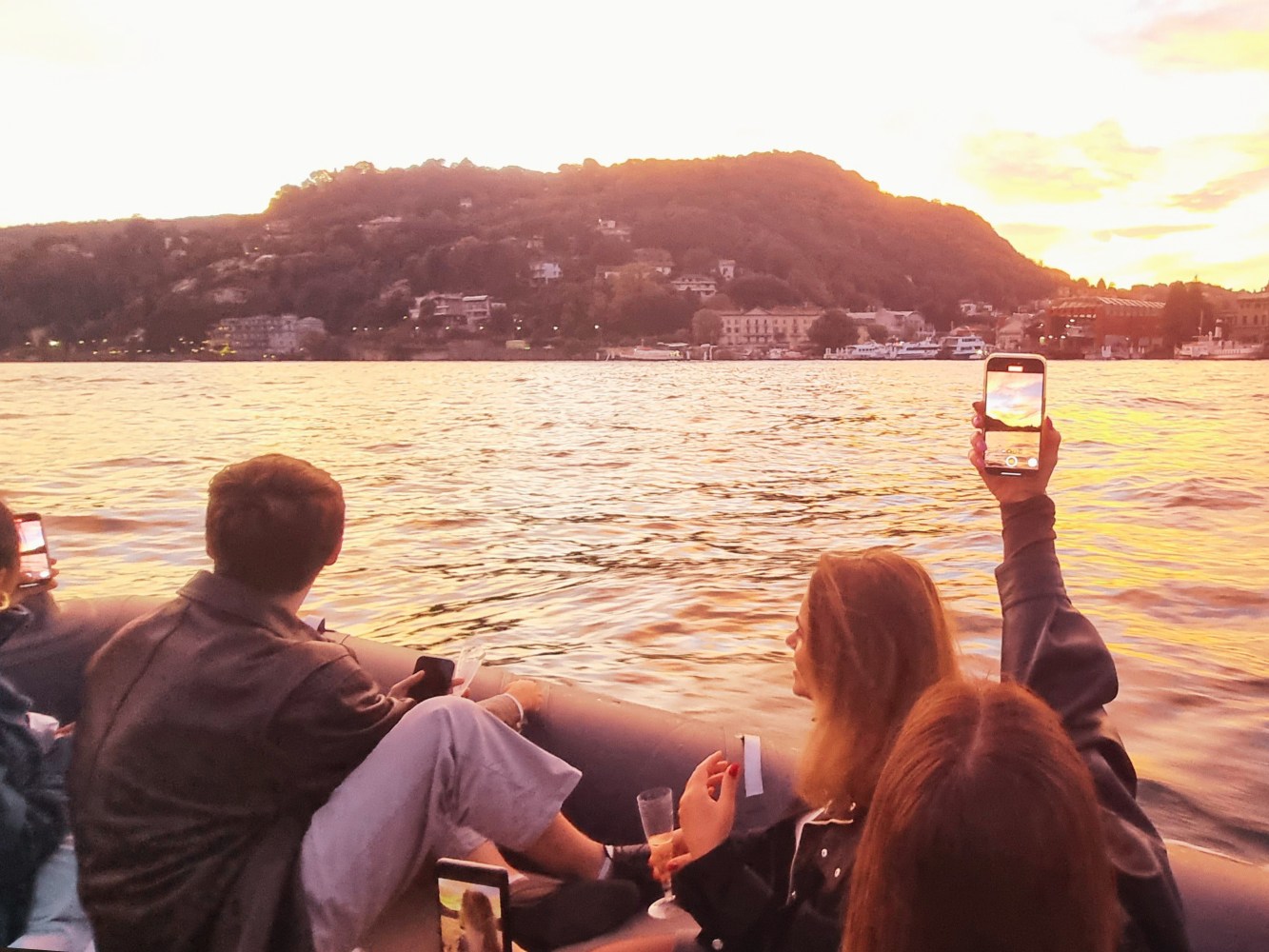 People on a boat at sunset, taking photos of a scenic view with hills in the background.