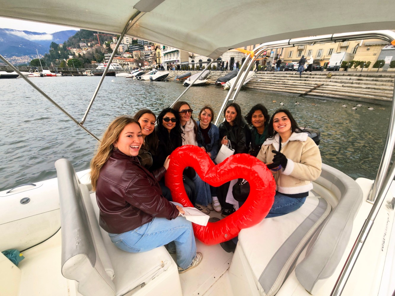 Group of women on a boat holding a red heart-shaped object with a scenic shoreline in the background.