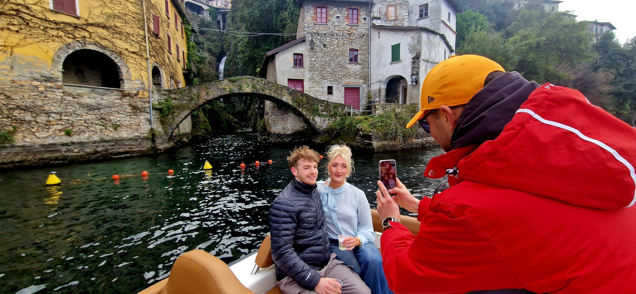 A person takes a photo of a couple sitting on a boat near historic buildings and an arched stone bridge.